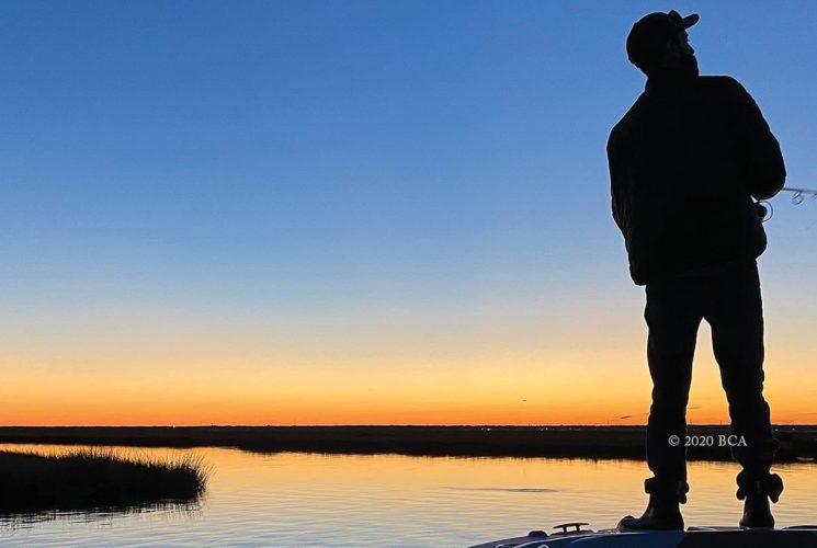 Man standing on boat fishing with water and land in the background at dusk