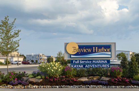 Bayview Harbor sign surrounded by pink, purple, and yellow flowers, green bushes, small tree, and rocks