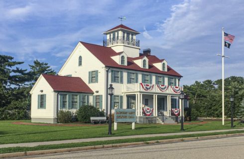 Exterior view of historic building painted white with sage green shutters and red roof and surrounded by green grass, bushes, and trees