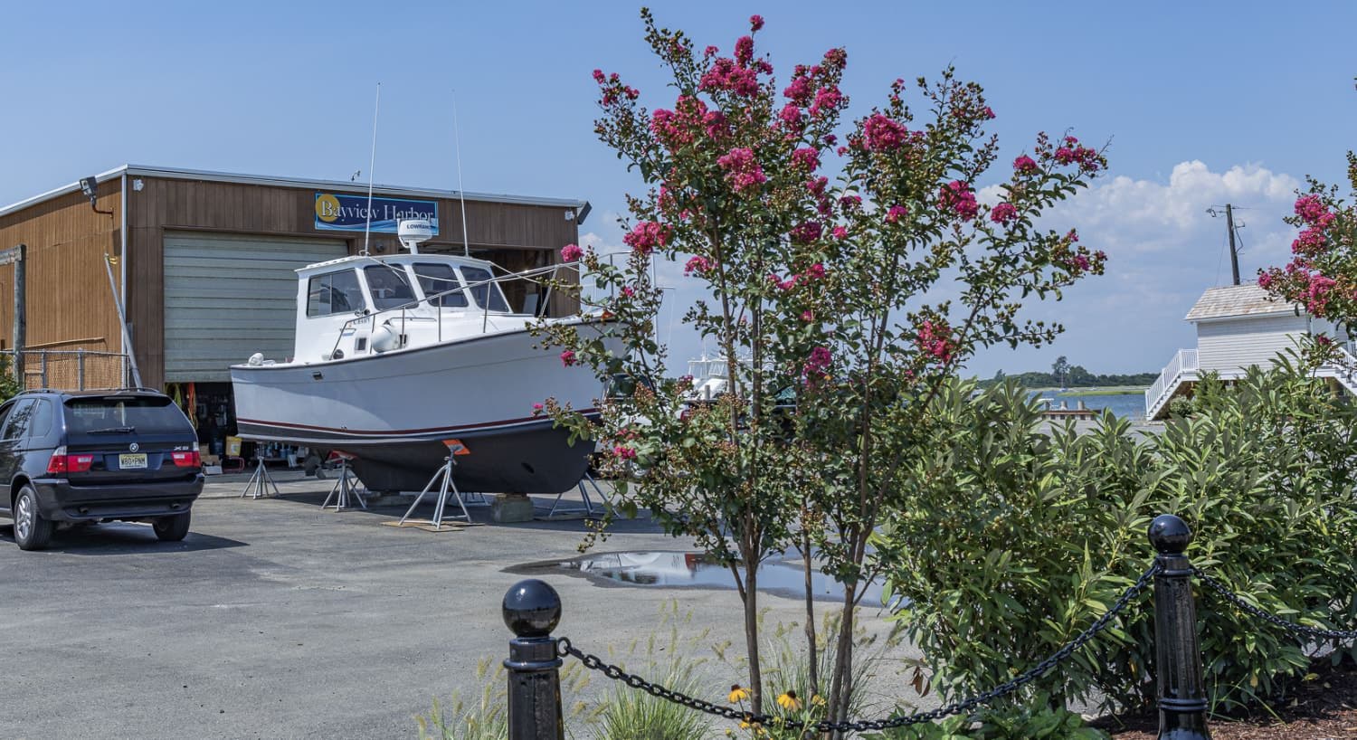 White boat out of the water in front of service building with small green bushes, flowers, and pink flowering trees nearby