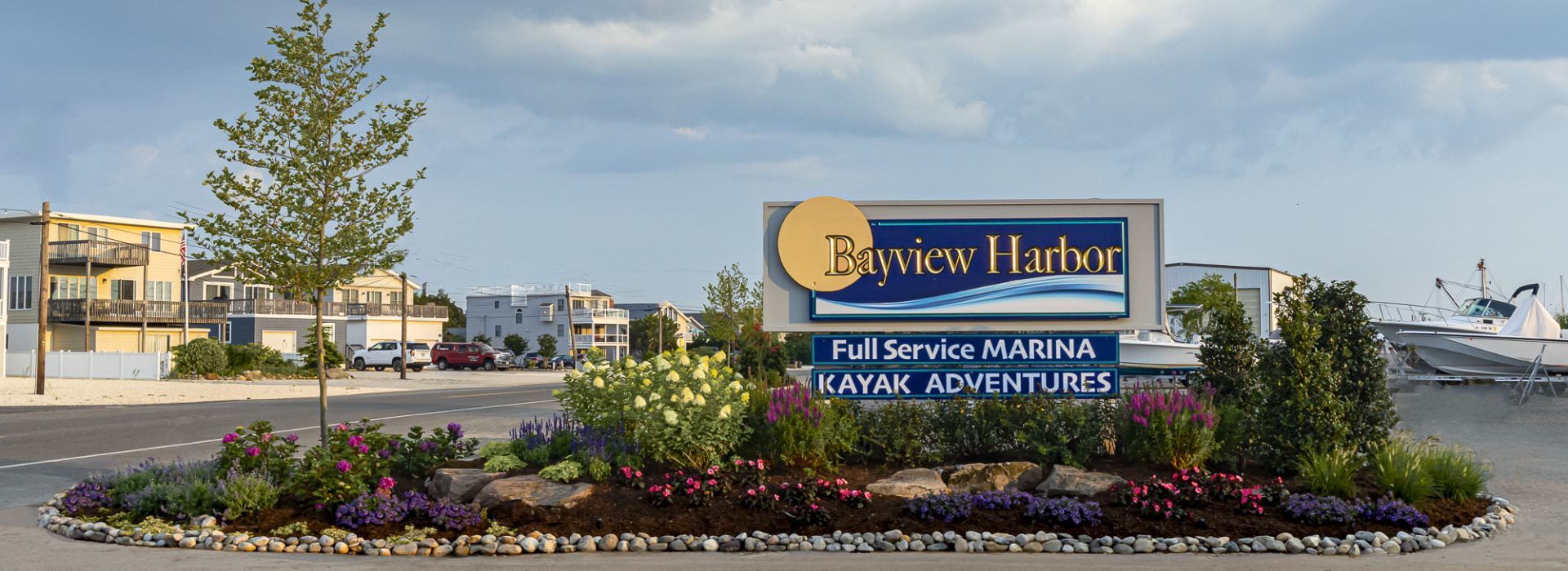 Bayview Harbor sign surrounded by pink, purple, and yellow flowers, green bushes, small tree, and rocks